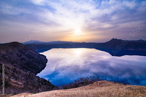 朝日の輝く空を湖面に映す山間の湖。北海道の摩周湖。