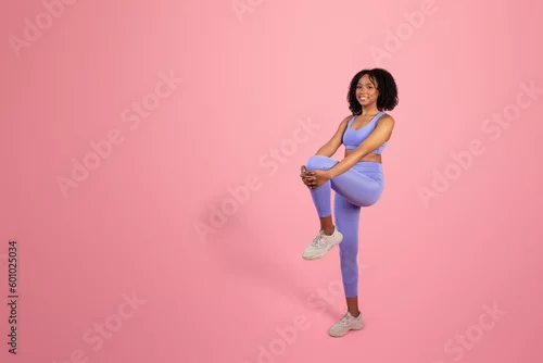 Obraz Cheerful millennial african american lady in sportswear doing leg warm up isolated on pink background