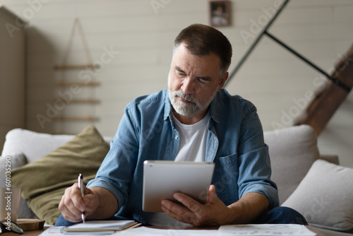 Cheerful senior man in casual clothing using digital tablet while sitting on the sofa at home.