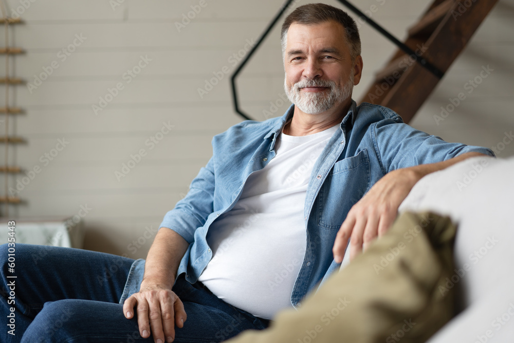 Fototapeta premium Happy mature man sitting on couch. Portrait of man relaxing at home with big smile.