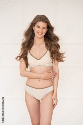 Confident beautiful young woman posing on white background at home in comfortable underwear smiling and looking away.