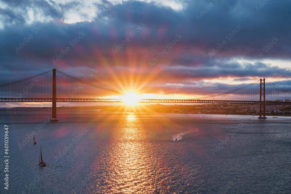 View of 25 de Abril Bridge famous tourist landmark of Lisbon connecting Lisboa and Almada on Setubal Peninsula over Tagus river with tourist yacht boats on sunset. Lisbon, Portugal View of 25 de Abril Bridge famous tourist landmark of Lisbon connecting Lisboa and Almada on Setubal Peninsula over Tagus river with tourist yacht boats on sunset. Lisbon, Portugal