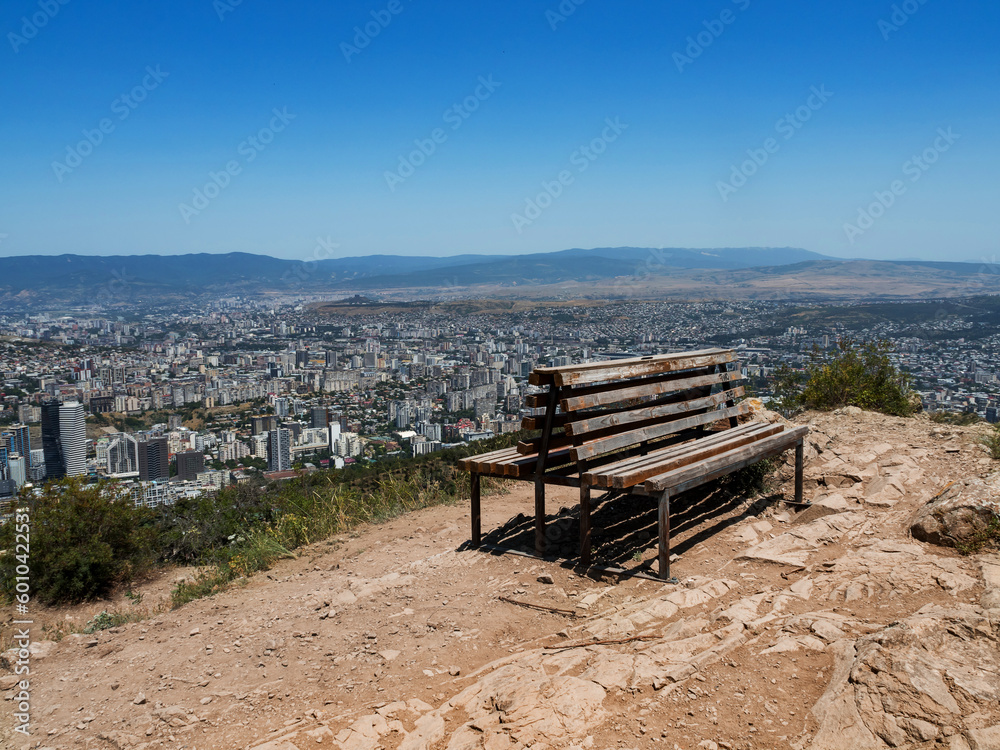 A double old wooden bench on top of a mountain with a beautiful view of the city in Georgia. Panorama of the city with a huge number of houses. Blue sky. Copy space. Place for text. No people A double old wooden bench on top of a mountain with a beautiful view of the city in Georgia. Panorama of the city with a huge number of houses. Blue sky. Copy space. Place for text. No people