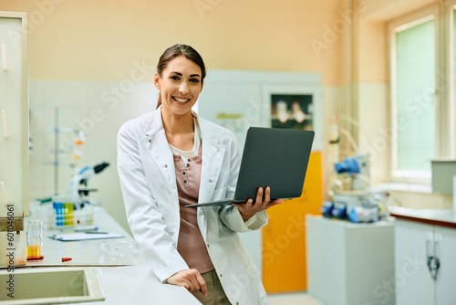 Happy female scientist working on laptop in laboratory and looking at camera.