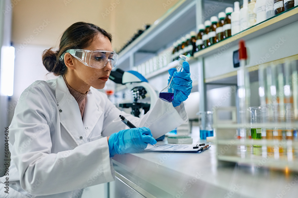 Female scientist taking notes while analyzing blue chemical liquid in lab.