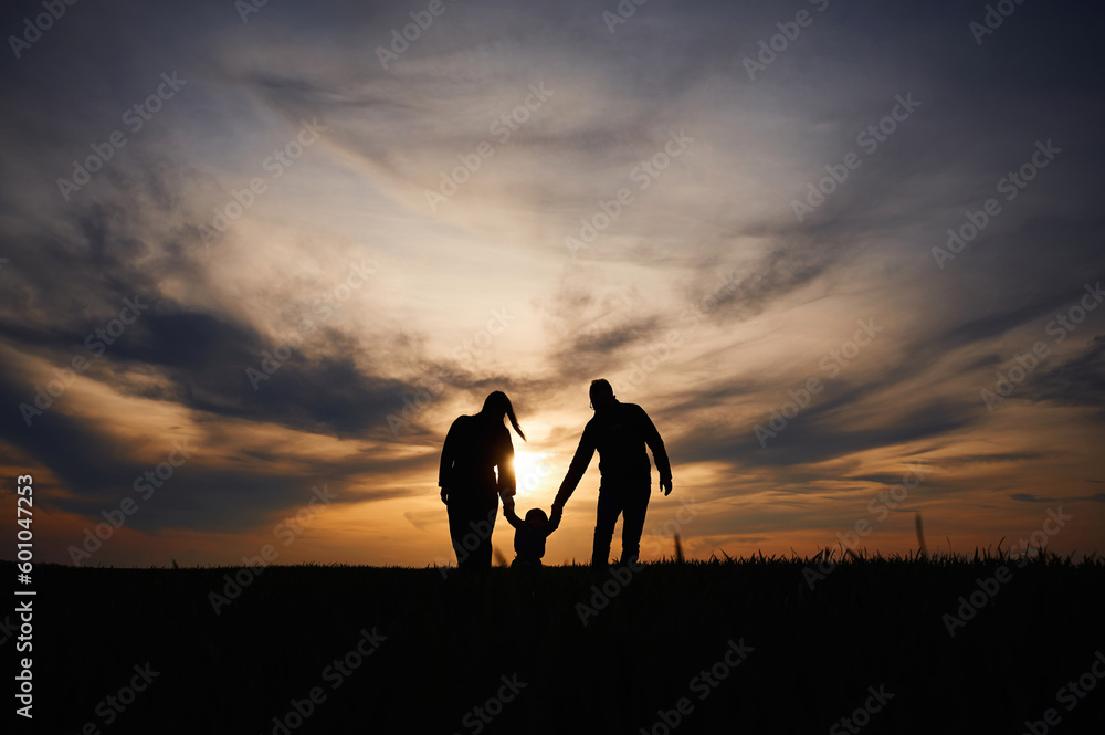 Silhouettes of father, mother and little baby son is outdoors against sunset dramatic sky in the field