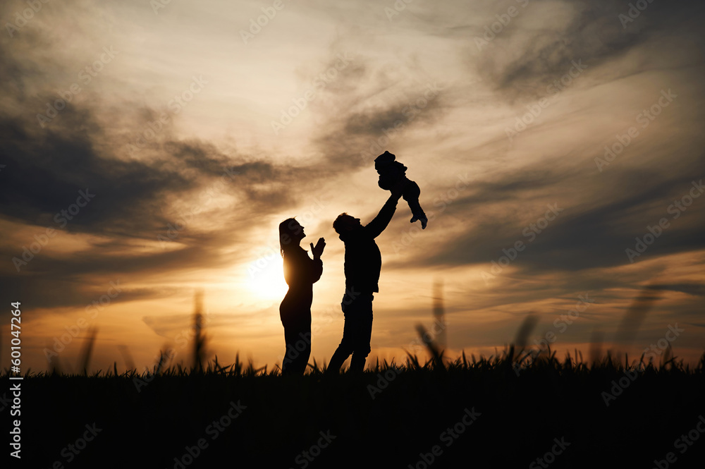 Holding little baby in hands. Silhouettes of father and mother that are outdoors against sunset dramatic sky in the field