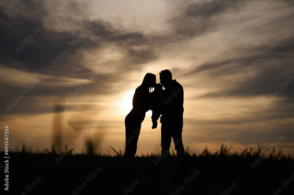 Silhouettes of father, mother and little baby son is outdoors against sunset dramatic sky in the field