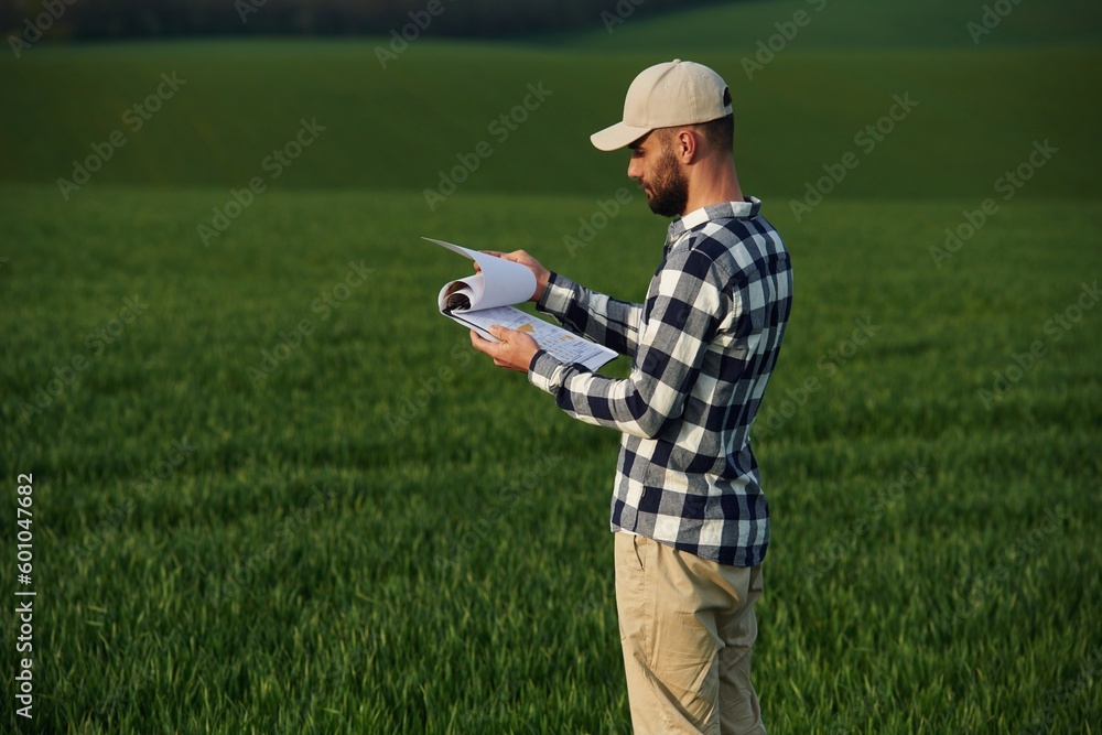 In checkered shirt and with notepad. Handsome young man is on agricultural field