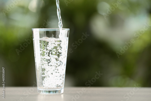 Pouring water into glass on white table against blurred green background, closeup. Space for text