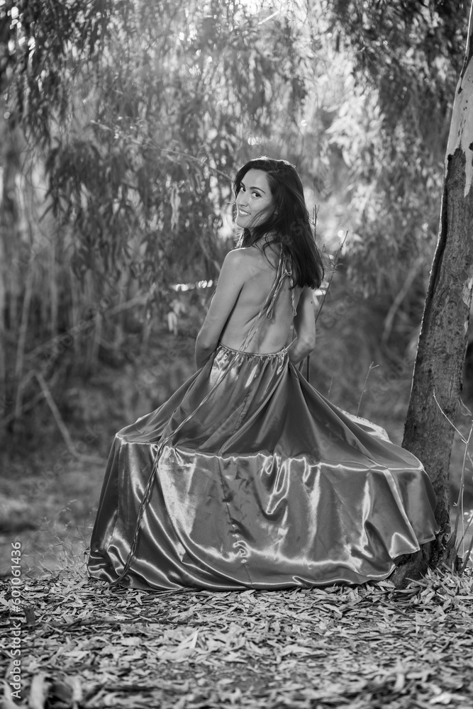 attractive brunette girl in the forest sitting on the log of a tree with her back to the camera, wearing a dress covering the surface of the trunk and revealing her back.
