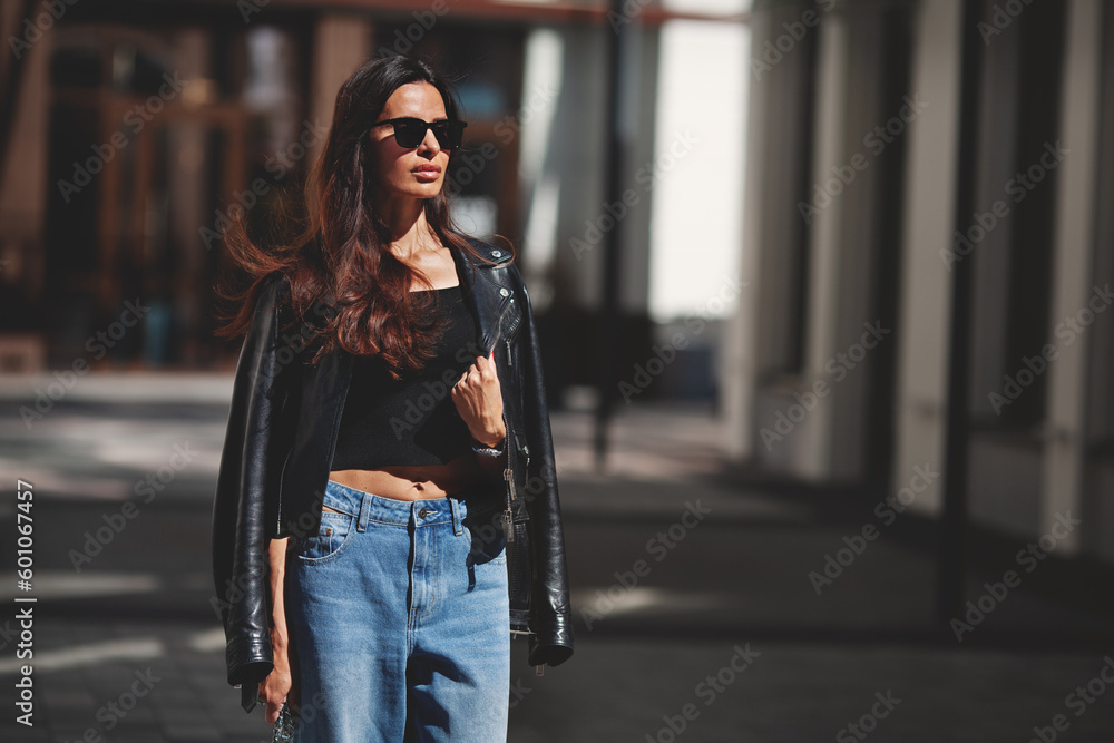Portrait of attractive fashionable brunette woman standing city street. Female model fashionista wears black leather jacket, jeans, short top, sunglasses. Street style