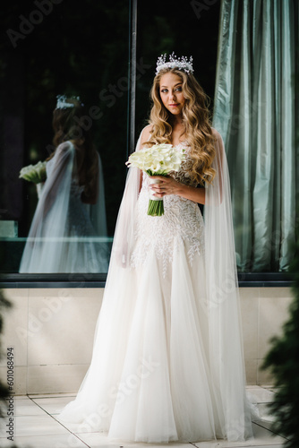 Young beautiful bride in elegant dress is standing and holding hand bouquet of white flowers and greens with ribbon. The girl holds a wedding bouquet outdoors.