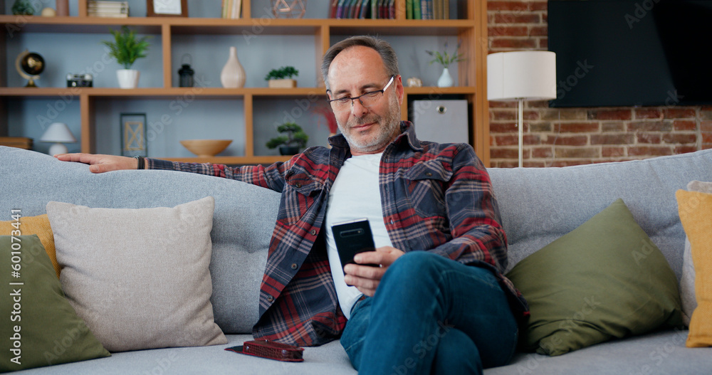 Attractive smiling adult bearded man in glasses relaxing on soft couch at home and uses his phone distracted to look out to window for a moment