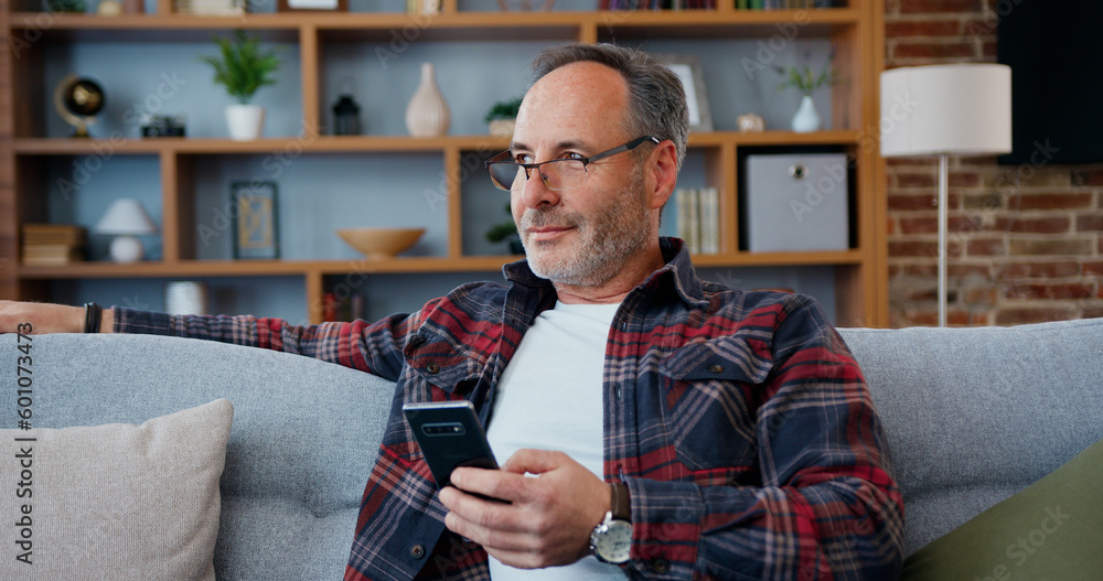 Handsome thoughtful happy bearded man in casual clothes sitting on the couch in living-room,looking out to window and then typing message on smartphone Handsome thoughtful happy bearded man in casual clothes sitting on the couch in living-room,looking out to window and then typing message on smartphone