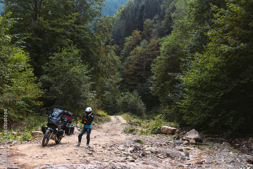 Biker girl in special motorcycle outfit, protection, knee pads and turtleneck. White helmet. Stands next to a long-distance touring motorcycle. Side bags and luggage. mountain road. Copy space.