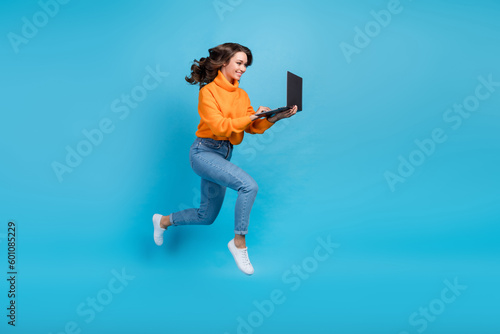 Full length photo of charming pretty lady dressed orange pullover jumping writing emails modern gadget isolated blue color background