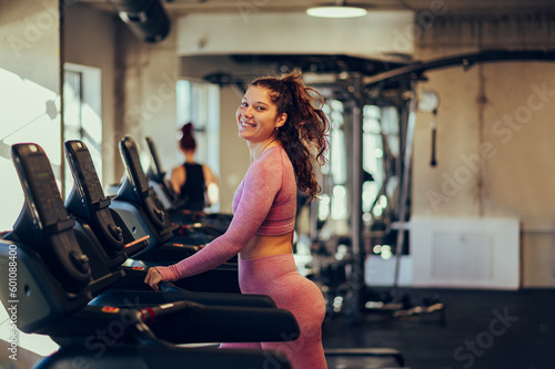 A happy chubby young sportswoman is walking on a treadmill in a gym and smiling at the camera.