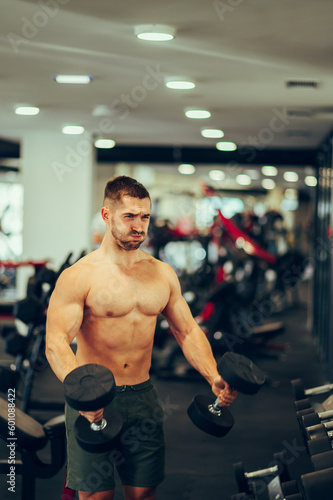 A strong fit shirtless weightlifter is doing hard workouts with a dumbbells with effort at the gym.