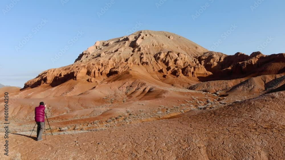A photographer takes a picture of unusual red rocks in the early morning. A man with a camera on a tripod, in a red down jacket. Rocky cliffs of sandstone red. Azhirzhar Tract, Kazakhstan A photographer takes a picture of unusual red rocks in the early morning. A man with a camera on a tripod, in a red down jacket. Rocky cliffs of sandstone red. Azhirzhar Tract, Kazakhstan