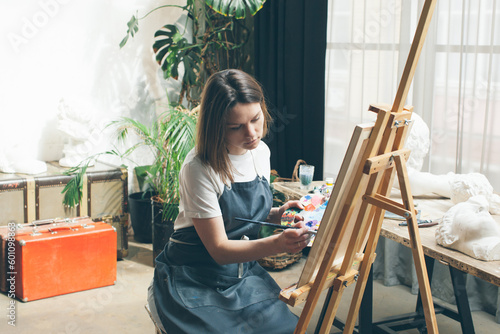 The artist paints in oils at the easel. In the hands of a palette and brush. White T-shirt and apron. Portrait. Creative person concept. Plants in the background. Workshop interior. Panoramic window.