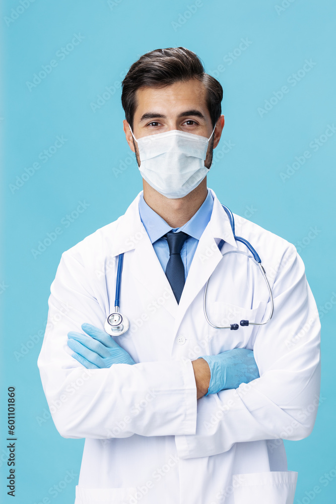 A male doctor in a white coat and a medical mask stands with his hands and looks at the camera on a blue isolated background, copy space, space for text