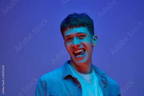 Portrait of young cheerful guy in casual shirt posing, smiling against gradient purple background in neon light. Feeling extremely happy. Concept of human emotions, lifestyle, youth, joy, fun