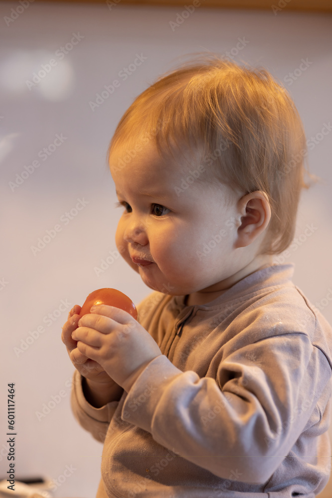 Baby in beige jumpsuit sitting on the table in the home kitchen and eating tomato Baby in beige jumpsuit sitting on the table in the home kitchen and eating tomato