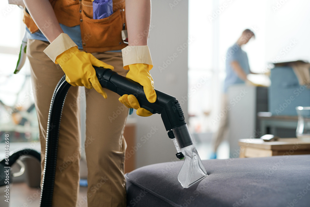 Close-up of cleaning service worker vacuuming furniture in apartment with her colleague wiping dust in background