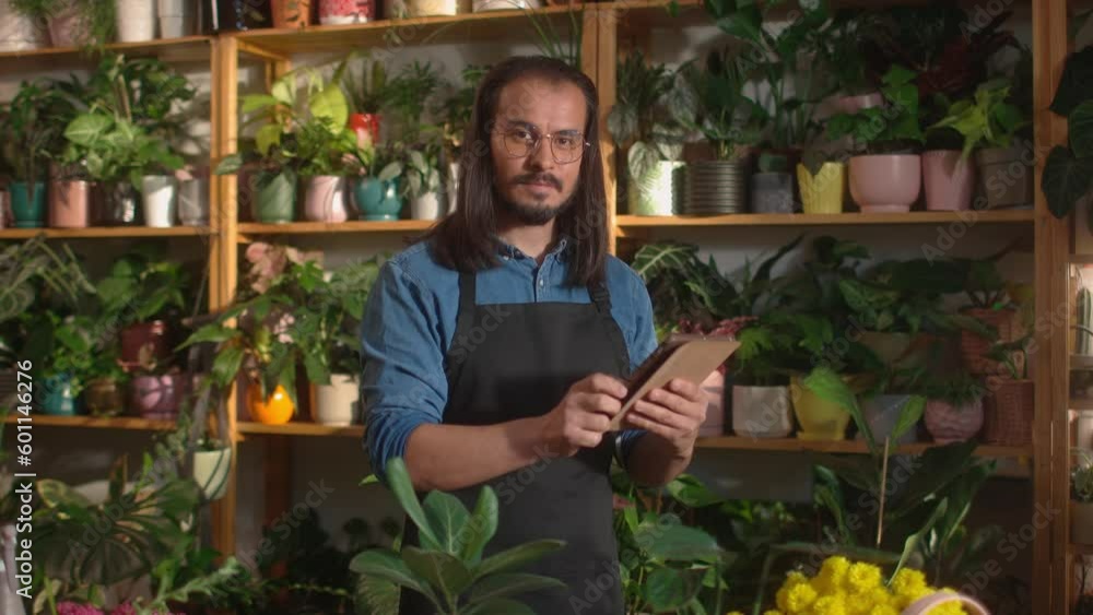 Attractive Caucasian gardener with apron standing behind table with greenery and flower pots. Adult man using tablet computer and tapping on sensor screen. Seriously looking straight into camera. Attractive Caucasian gardener with apron standing behind table with greenery and flower pots. Adult man using tablet computer and tapping on sensor screen. Seriously looking straight into camera.