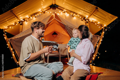 Happy family with lovely baby relaxing and spend time together in glamping on summer evening near cozy bonfire. Luxury camping tent for outdoor recreation and recreation. Lifestyle concept