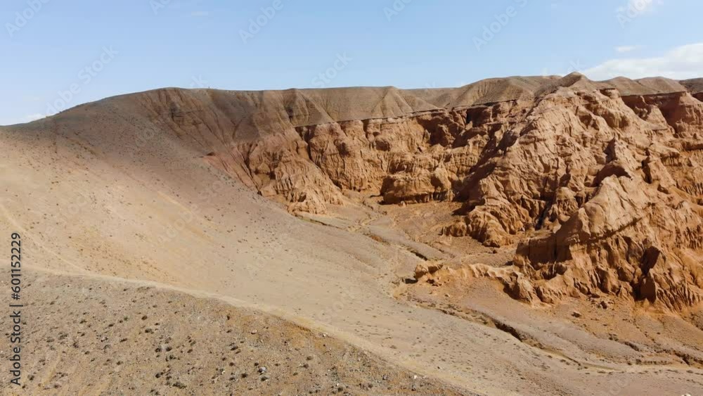 The photographer walks along a hill among unusual red rocky mountains. A man with a camera on a tripod, in a lemon windbreaker. Rocky cliffs of red sandstone. Tract Azhirzhar, Kazakhstan The photographer walks along a hill among unusual red rocky mountains. A man with a camera on a tripod, in a lemon windbreaker. Rocky cliffs of red sandstone. Tract Azhirzhar, Kazakhstan