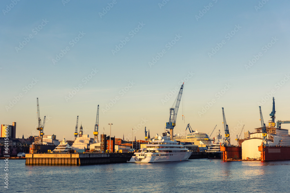 Scenic panoramic view large modern luxury super yacht at maintenance service against construction cargo cranes at dry dock shipyard in Hamburg port sunset sky. Big vessel manufactoring site industry