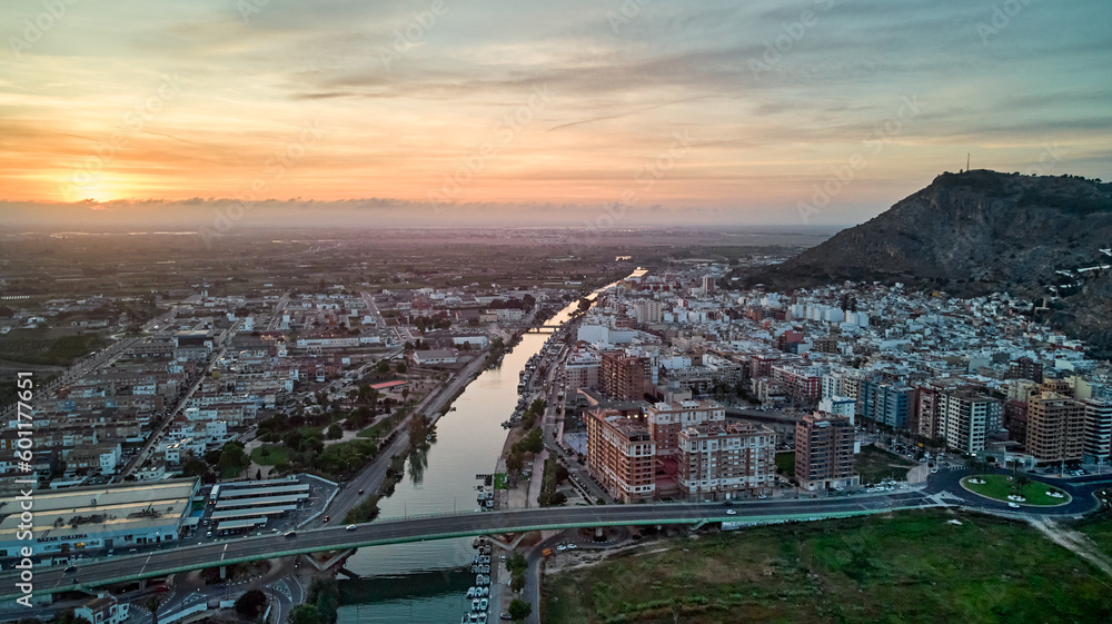 Sunset over city, buildings stretch to horizon with mountains and river on either side, bridge spans river, warm orange glow creates serene atmosphere.