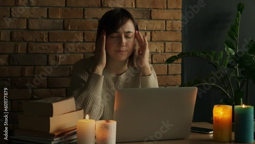 Caucasian woman is under stress using laptop at home during electricity outage or blackout with lit candles. Tired woman rubbing her temples and having a headache