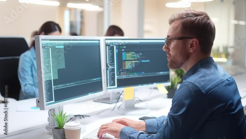 Hardworking Caucasian programmer sitting in front of multiple displays. Young man writing code for creating app or program on website. Working in IT-company. In background visible workers at computers