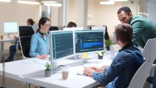 Mixed-raced IT-programmers speaking about business project in office. Hardworking man sitting on work chair and typing programming code on keyboard. Positive people looking at monitors and laughting.