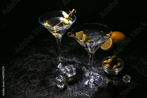 Glasses of tasty martini, ice cubes, green olives and lemon on dark background