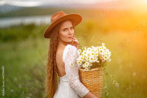 A middle-aged woman in a white dress and brown hat stands on a green field and holds a basket in her hands with a large bouquet of daisies. In the background there are mountains and a lake.