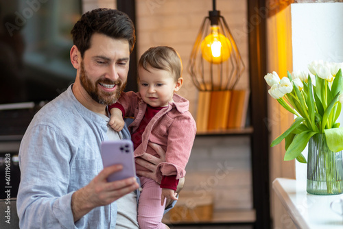 Bearded father standing with baby daughter and mobile phone having video call or selfie