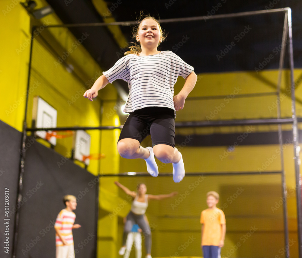 Full length of excited casual schoolchild girl having fun while jumping high on colorful trampoline at game club