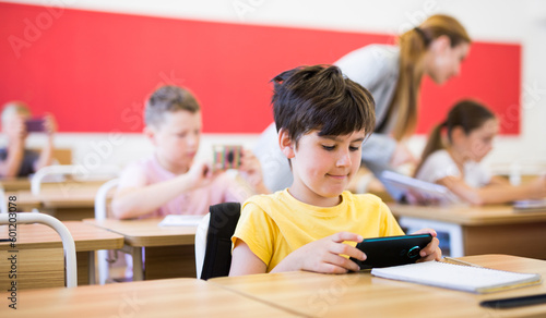 Portrait of schoolboy sitting at desk and using mobile phone during lesson in elementary school