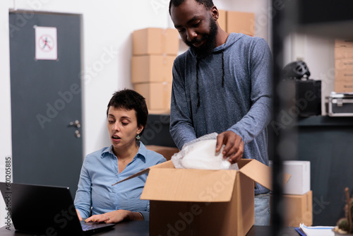 Storehouse worker putting client order in carton box wrapping with bubble wrap while supervisior woman checking transportation detalies on laptop computer. Warehouse job concept photo