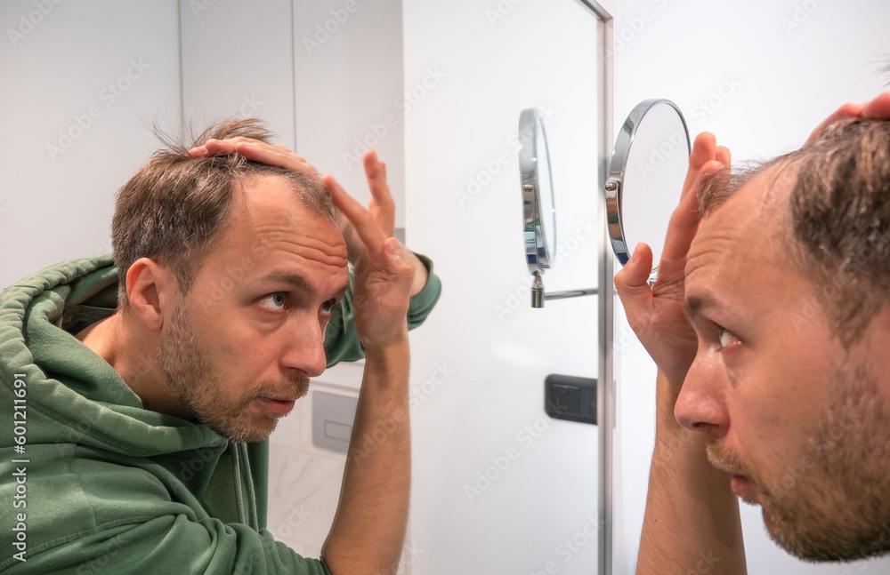 Balding middle age man looks in mirror and checks how much hair he lost. - obrazy, fototapety, plakaty 