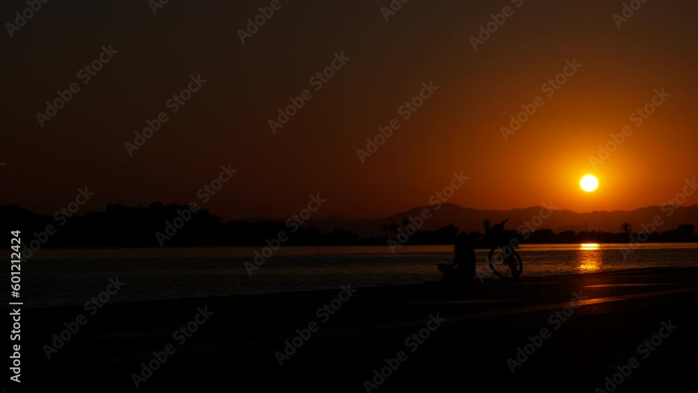 Woman at sunset with a bicycle near the water. Silhouette of a woman with a bicycle sitting on the sea embankment against the backdrop of sunset. Woman at sunset with a bicycle near the water. Silhouette of a woman with a bicycle sitting on the sea embankment against the backdrop of sunset.