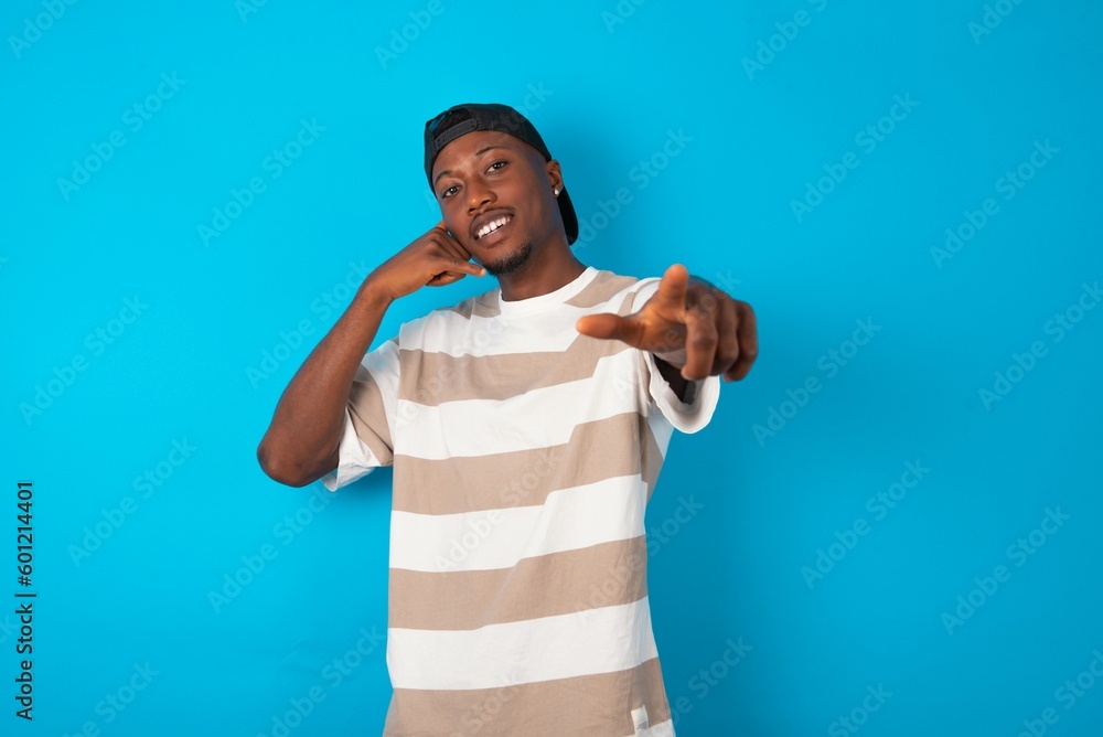 Naklejka premium Handsome man wearing striped t-shirt and cap over blue background smiling cheerfully and pointing to camera while making a call you later gesture, talking on phone
