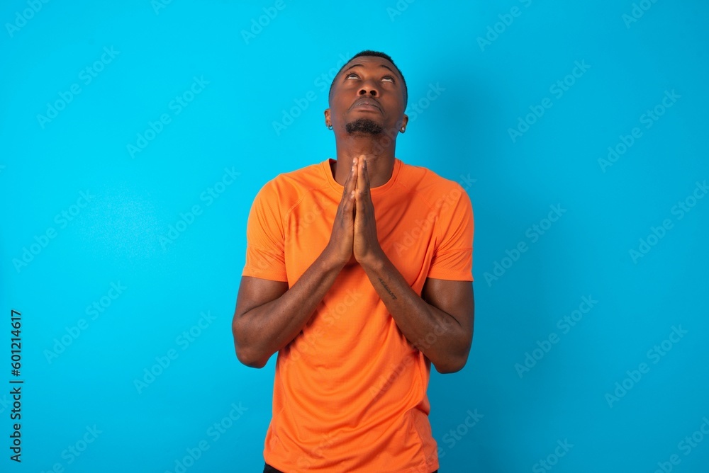 Handsome man wearing orange T-shirt over blue background begging and praying with hands together with hope expression on face very emotional and worried. Please God Handsome man wearing orange T-shirt over blue background begging and praying with hands together with hope expression on face very emotional and worried. Please God