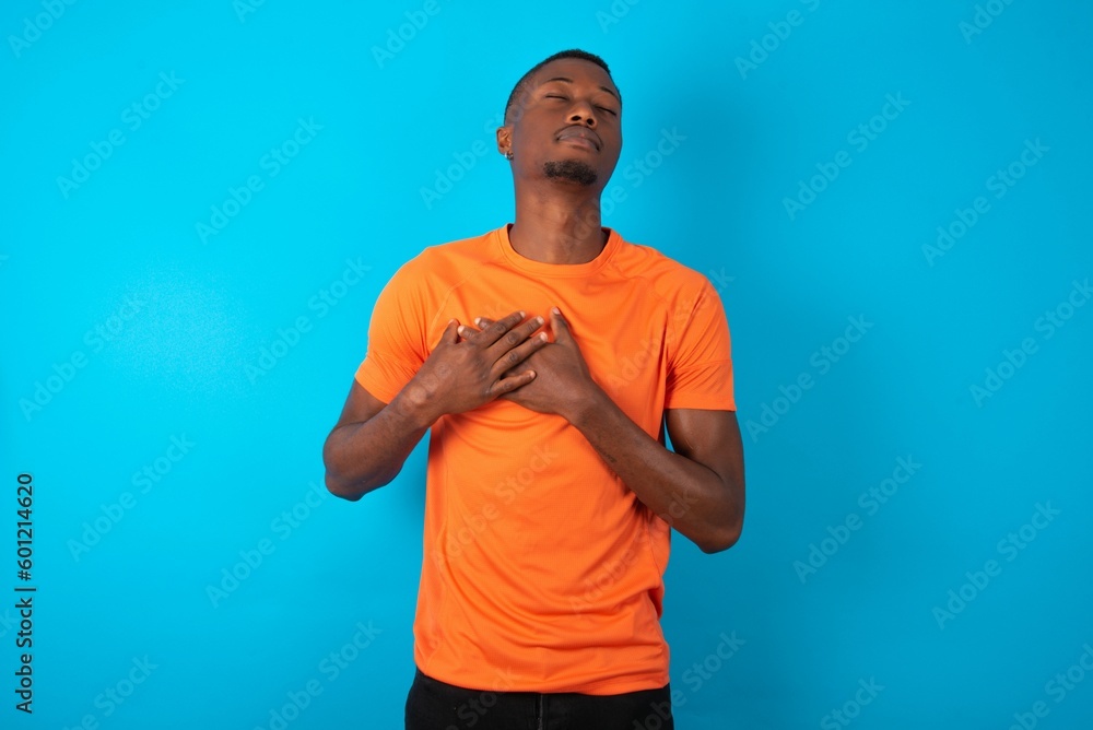 Handsome man wearing orange T-shirt over blue background smiling with hands on chest with closed eyes and grateful gesture on face. Health concept. Handsome man wearing orange T-shirt over blue background smiling with hands on chest with closed eyes and grateful gesture on face. Health concept.