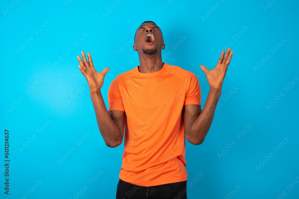 Handsome man wearing orange T-shirt over blue background crazy and mad shouting and yelling with aggressive expression and arms raised. Frustration concept. Handsome man wearing orange T-shirt over blue background crazy and mad shouting and yelling with aggressive expression and arms raised. Frustration concept.