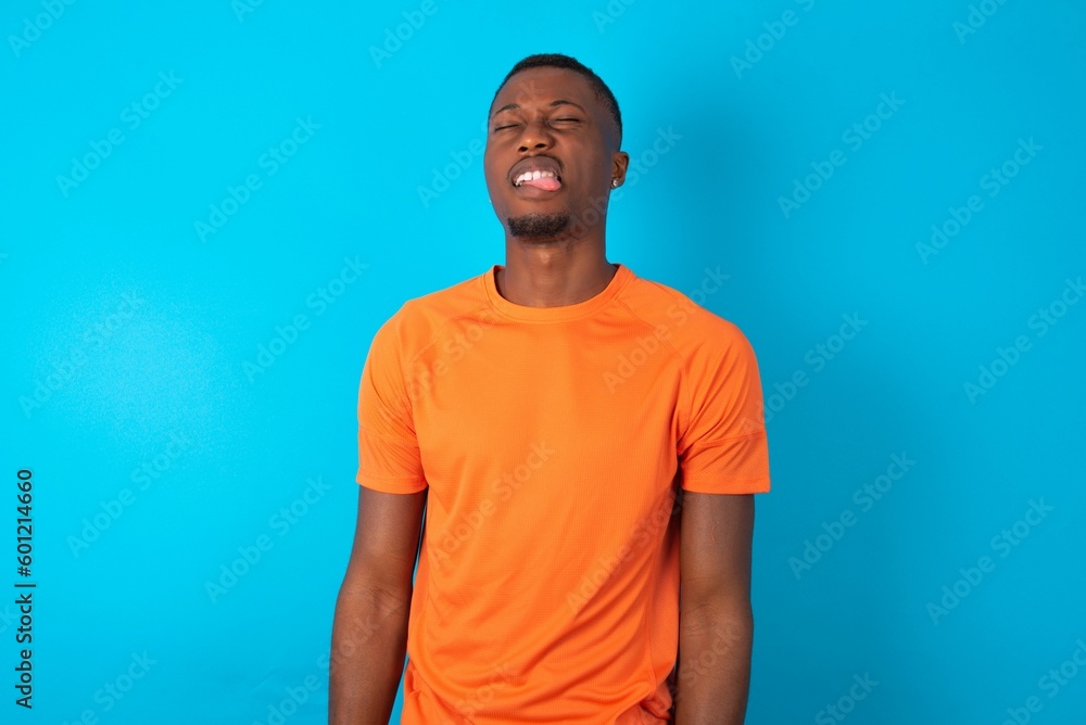 Handsome man wearing orange T-shirt over blue background sticking tongue out happy with funny expression. Emotion concept. Handsome man wearing orange T-shirt over blue background sticking tongue out happy with funny expression. Emotion concept.